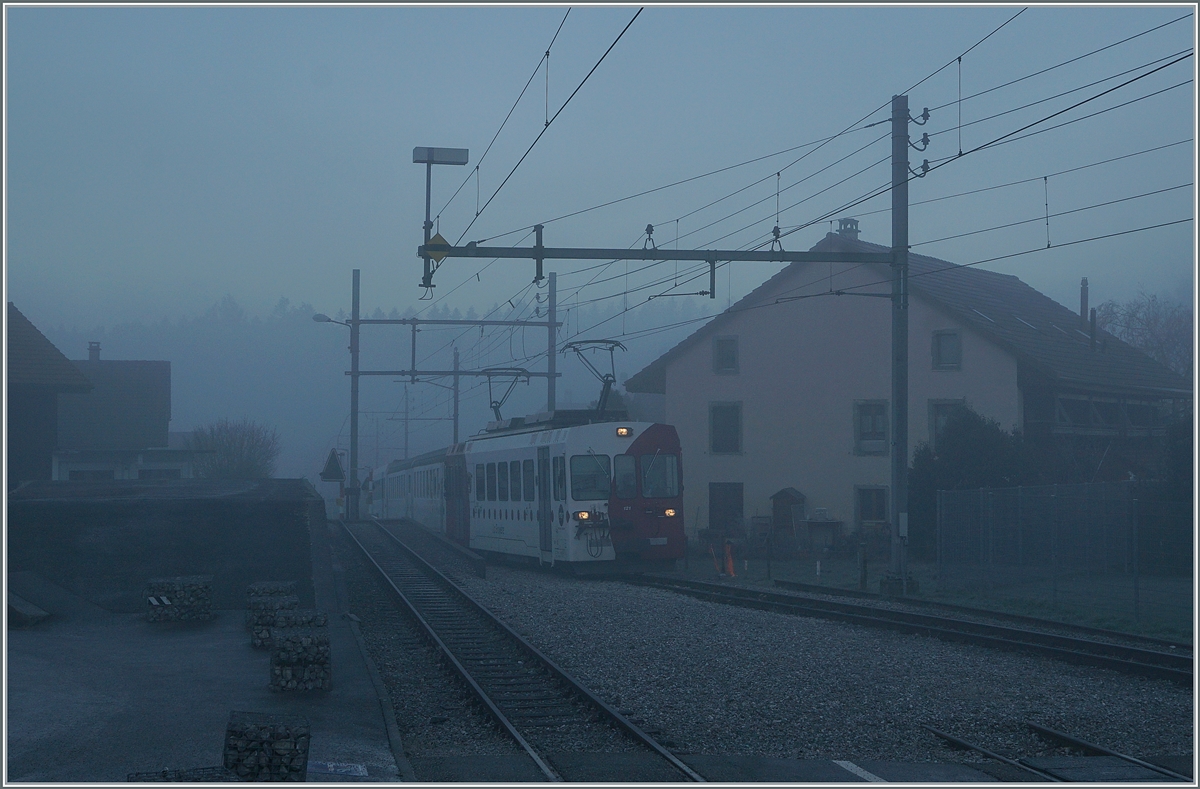 Bei dichtem Nebel und noch in der Morgendämmerung erreicht der TPF Be 4/4 121 mit dem B 207 (ex MOB), B209 (ex MOB) und dem ABt 221 den Bahnhof von Broc Village. Der Zug ist als RER 60 14954 von Bulle nach Broc Farbrique unterwegs. 

26. Nov. 2020