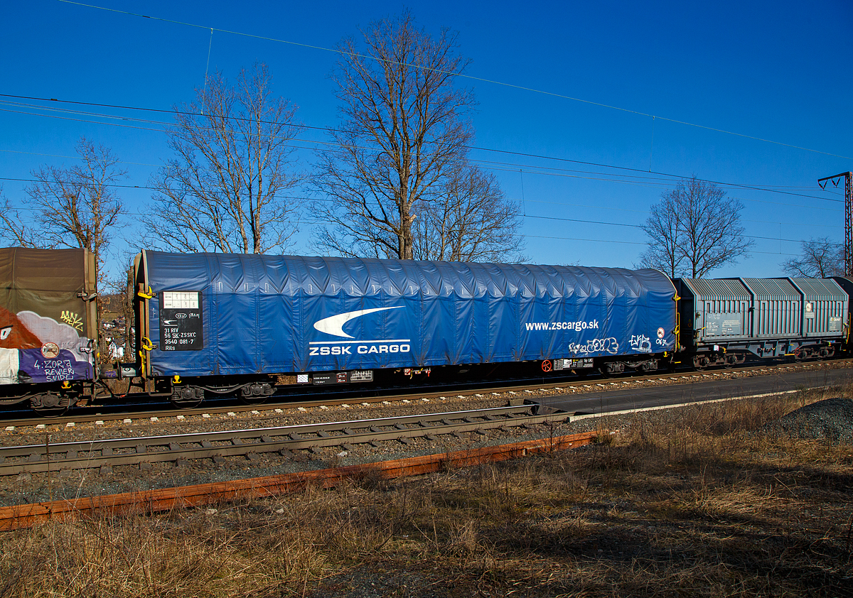 4-achsiger Drehgestell-Flachwagen mit verschiebbarem Planenverdeck, 31 56 3540 081-7 SK-ZSSKC, der Gattung Rils, der slowakischen ZSSK Cargo (Železničná spoločnosť Cargo Slovakia), am 10.03.2022 im Zugverband bei einer Zugdurchfahrt in Rudersdorf (Kr. Siegen).

Die Ausrüstung mit Planen-(Schnell-)Verdeck ermöglicht den Transport von Gütern, für die ein Nässeschutz erforderlich ist. Das Verdeck zeichnet sich durch einfache Handhabung mit Ein-Mann-Bedienung, Leichtgängigkeit und Zentralverriegelung aus. 

Das Planendach wird in geschlossener Stellung manuell verriegelt. Die 4-Punkt-Zentralverriegelung an der Stirnwand ist robust und einfach. Durch die gewählte Anordnung der Betätigungseinrichtung ist das Entriegeln des Planendaches sowohl vom Erdboden als auch von einer Rampe aus möglich. Die beidseitig an jeder Stirnwand angeordneten Verriegelungshaken einschließlich ihrer Betätigungshebel sind über eine drehbar gelagerte Welle miteinander verbunden. Hierdurch wird das Betätigen von nur einer Wagenseite möglich.
Das Planendach lässt sich jeweils zu einem Wagenende hin so weit zusammenschieben, dass jeweils ca. 2/3 des Wagens freigelegt werden kann. 

Die Wagenabdeckung erfolgt mit einer PVC-beschichteten Gewebeplane, die auf 18 Rohrrahmen aufliegt und über Laufwagen in Wagenlängsrichtung verschoben werden kann. Die unteren Laufwagenrollen wirken als Abhebesicherungen. Sie greifen unter die Laufschienenunterkanten und verhindern so ein Abheben der Hauben bei Auflaufstößen oder seitlicher Windbelastung.

TECHNISCHE DATEN:
Spurweite: 1.435 mm (Normalspur)
Anzahl der Achsen: 4 in 2 Drehgestellen
Länge über Puffer: 19.900 mm
Drehzapfenabstand:  14.860 mm
Achsabstand in den Drehgestellen: 1.800 mm
Eigengewicht: 24.360 kg
Ladelänge: 18.500 mm
Ladefläche: 49,3 m²
Maximale Ladegewicht: 55,7 t (Streckenklasse C)
Höchstgeschwindigkeit: 100 km/h (beladen) / 120 km/h (leer)
Kleinster befahrbarer Gleisbogenhalbmesser: R 35 m
Feststellbremse: ja
Bremse: DK-GP (LL) SZ 12
Bremssohle: IB 116
Intern. Verwendungsfähigkeit:  RIV
