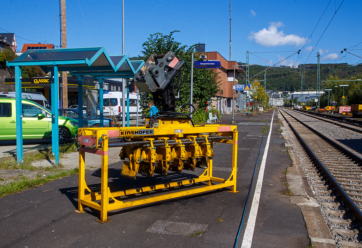 Bahnschwellenleger für Bagger vom Typ RUS05 im Geräteständer, der Joseph Hubert GmbH & Co. KG, am 30.09.2022 abgestellt am Bahnsteig vom Bahnhof Niederschelden (geografisch eigentlich Niederschelderhütte).

Der Hersteller (Baujahr 2020) von dem Bahnschwellenleger ist die KINSHOFER GmbH / RF SYSTEM in (Oberbayern).

Mit dem RUS05 Bahnschwellenleger können Betonschwellen mittig
gegriffen und dann einfach und effektiv positioniert werden, er ist geeignet für den Anbau an Bagger mit 14t bis 24t Dienstgewicht.

Technische Merkmale:
• Greifarme mit Synchronlauf und Untergreifen der Schwellen.
• Effizient: Mehrere Betonschwellen werden gleichzeitig durch hydraulische Greifer mittig geklemmt und anschließend auf das Schotterbett gelegt. Dies spart Zeit im Vergleich zur Einzelpositionierung der Schwellen.
• Sicher: Ein spezielles Halteventil garantiert ein sicheres Halten der Last, selbst bei Druckabfall.
• Variabler Greifbereich: Die Position der Greifarme ist innerhalb des vorgegebenen Bereichs verstellbar.
• Flexibilität: Für fast alle taillierten Bahnschwellentypen. Höhenanschlag je Schwelle einstellbar.
• Für enge Bereiche, da die Schwellen mittig gegriffen werden.
• Präzise Positionierung dank aufgebautem 10t-Drehmotor mit 4 Öldurchführungen und Schlauchschutz.
• Inklusive Geräteständer.
• Einstellbarer Schwellenabstand: 600mm / 620mm / 650mm

Der RUS05 Bahnschwellenleger ist ein hoch-spezialisiertes Gerät zum Greifer und Positionieren von Bahnschwellen nach einer bestimmten Anordnung. Der Abstand der Schwellen kann abhängig von den Anforderungen schnell und einfach angepasst werden. Die Bahnschwellen werden mittig von den voreingestellten, hydraulischen Greifarmen geklemmt und auf dem Schotterbett positioniert. Im Vergleich zur individuellen Positionierung der Bahnschwellen wird viel Zeit eingespart. So gut wie alle Arten von taillierten Bahnschwellen können sicher geklemmt werden, dank integriertem Lasthalteventil auch bei plötzlichem Druckabfall.

Der RUS05 wird inklusive praktischem Geräteständer ausgeliefert, zur einfachen Lagerung und Transport des Gerätes wenn nicht am Trägergerät montiert. Als Variante gibt es auch den RUS2 05 Bahnschwellenleger, dieser ist wie der RUS05 speziell für das Verlegen von fast allen Schwellentypen konzipiert, allerdings werden Bahnschwellen bei dieser Variante über die Enden geklemmt und nicht mittig. Beide Schwellenleger sind mit einem 10t-Drehmotor ausgestattet und können 5 Schwellen gleichzeitig greifen.

TECHNISCHE DATEN Bahnschwellenleger RUS05:
Greifbereich: 150 bis 265 mm
Gewicht: 1.300 kg
Länge: 2.820 mm
Höhe: 1.553 mm
Breite: 825 mm
Anzahl der Greifzangen: 5
Backenbreite: 330 mm (Schwellenbreite 300 / 320 mm)
Max. Gewicht  Einzelschwelle 350 kg (5 x 350 = 1.750 kg Gesamtlast)