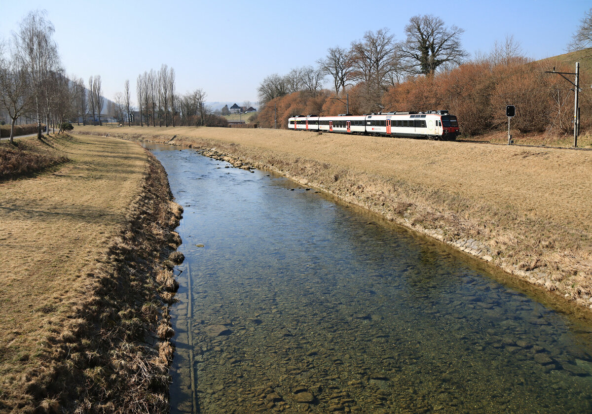Bei Moudon, im Talboden der Broye, jetzt im Canton de Vaud. Ein Domino-Zug der S8 Palézieux-Payerne fährt dem schön eingebetteten Fluss entlang. 10.März 2022  