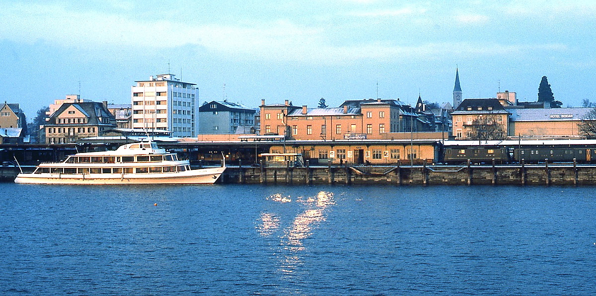 Blick von der frühmorgendlichen Bodenseefähre aus Friedrichshafen auf den Bahnhof Romanshorn. Rechts fährt gerade eine Re 4/4 mit einem Personenzug ein. Das Foto entstand an einem Februartag Anfang der 1980er Jahre.