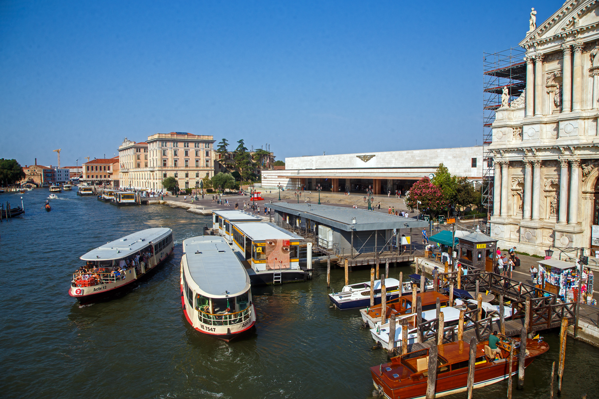 Blick der über den Canal Grande überspannenden Scalzi-Brücke (Ponte degli Scalzi) auf das Empfangsgebäude vom Bahnhof Venezia Santa Lucia am 24.07.2022. 

Der Bahnhof Venezia Santa Lucia der Hauptbahnhof von Venedig. Er ist ein Kopfbahnhof und der einzige Bahnhof der Altstadt von Venedig. Er liegt auf der Insel San Geremia im Sestiere Cannaregio. Seinen Namen hat er von der Santa-Lucia-Kirche, die ursprünglich an diesem Ort stand und für den Bau des Bahnhofs 1861 abgerissen wurde. Ein Gedenkstein auf der Mitte des Platzes erinnert an die ehemalige Kirche.

Neben der Brücke bzw. vor dem Bahnhof liegt die Haltestelle Ferrovia die von Vaporetto (Wasserbuse) und Motoscafi (Wasser-Taxi) bedient werden. An der Haltestelle halten Linien ACTV. Unweit (400 m) in südlicher Richtung (hier im Bild nach links) der Piazzale Roma mit dem Omnibusbahnhof und der Straßenbahnhaltestelle. 

Rechts nur etwas angeschnitten die Scalzi-Kirche (Santa Maria di Nazareth, umgangssprachlich Scalzi), nach der die Brücke ihren Namen bekommen hat.
