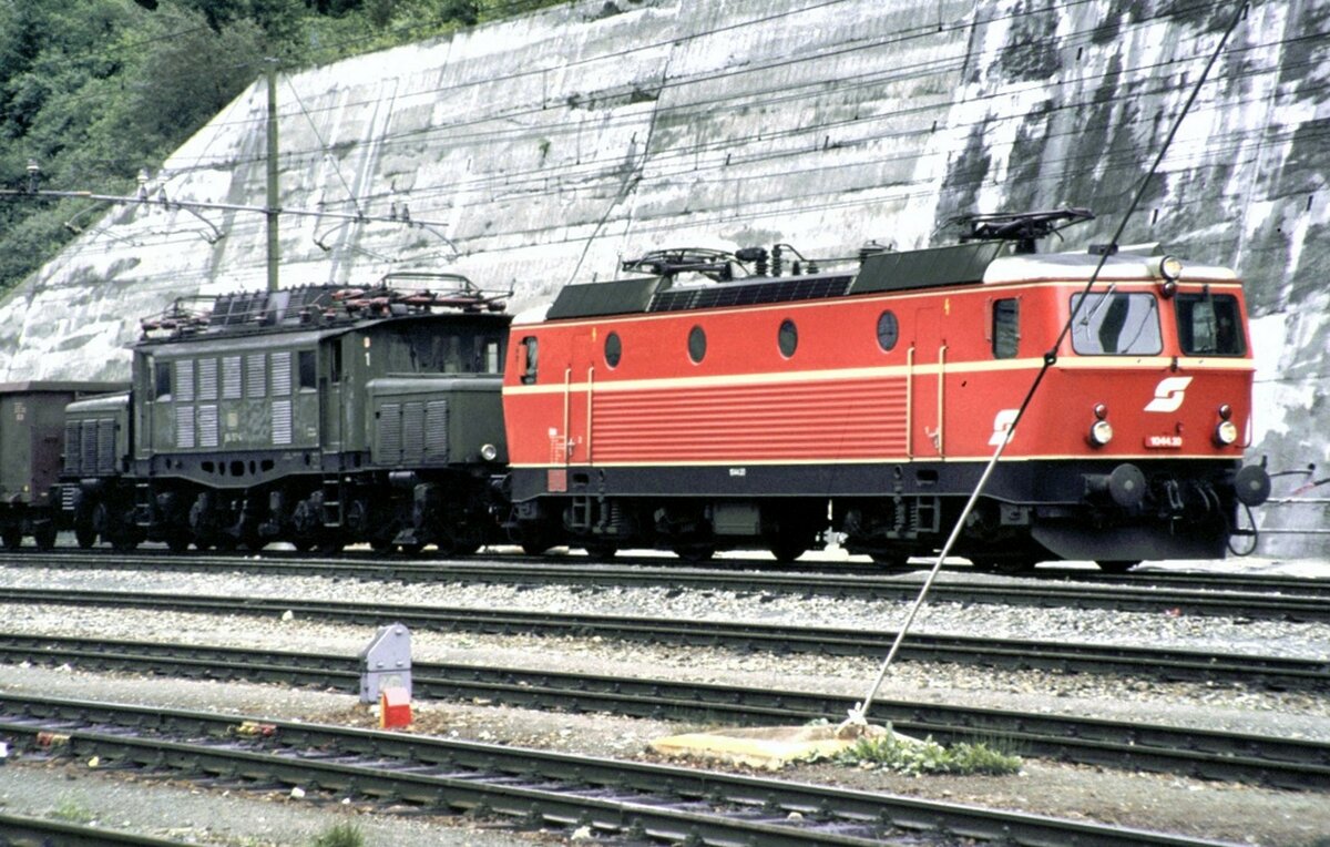 DB 194 und ÖBB 1044 sind mit ihremGüterzug an der Brenner Station abgebügelt in den Gleichstrombereich gerollt. Bald kommt eine FS Diesellok und zieht bzw. schiebt die Loks wieder in den Wechselstrombereich für die Rückfahrt am 01.10.1982. Heute ist alles anders geregelt und wird durch automatische Schaltungen gesteuert.