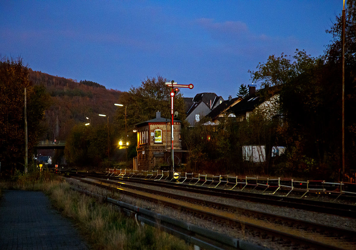 
Ein abendlicher Blick auf das 1901 gebaute Wärterhebelstellwerk Herdorf Ost (Ho) am 05.11.2020. 

Davor auf Gleis 2 das (Flügelsignal) Formhauptsignal, der obere Flügel zeigt waagerecht nach rechts und das obere Licht ist „rot“ und somit zeigt es Hp 0 (Halt) an. Unten vor dem Hauptsignal das Schutz- und Gleissperrsignal (waagerechter schwarzer Streifen auf runder weißer Scheibe), welches somit Sh 0 (Halt! Fahrverbot)  anzeigt.

Weit hinten kurz vor der Brücke sieht man das Signal Sh 2 – „Schutzhalt.“, die rechteckige rote Scheibe mit weißem Rand und davor das rote Licht. Eine Weiterfahrt ist somit auf der Hellertalbahn (KBS 462) in Richtung Neunkirchen nicht möglich, da noch Arbeiten ausgeführt werden.
