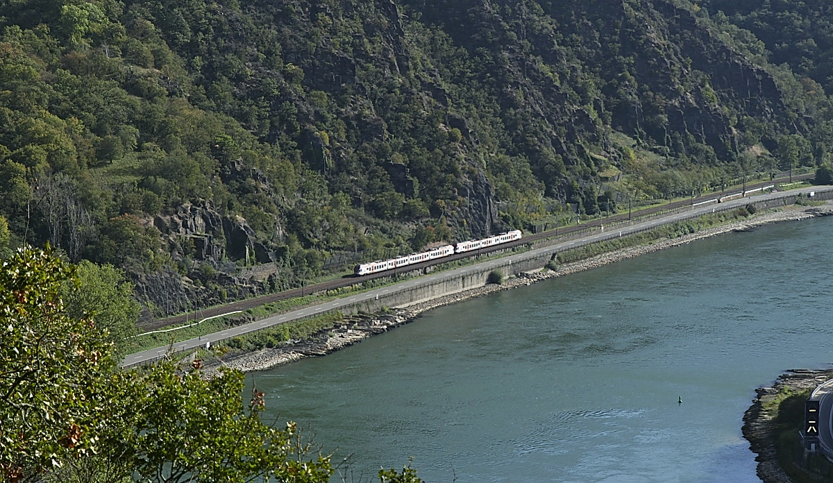 Ein Blick vom Loreleyfelsen nach Süden mit einem vier- und dreiteiligen Flirt von VIAS, der am 11.10.2023 als Rheingau-Linie RB 10 von Frankfurt am Main nach Neuwied unterwegs ist. Im Gegensatz zur rechtsrheinischen Bahn mit dem dichten ICE-, IC/EC- und RE-Verkehr ist dies die einzige Linie, die im Mittelrheintal auf der linken Rheinseite regelmässig verkehrt. An der Loreley liegt die engste Stelle des Mittelrheintals, hier und im Oberlauf ist der Fluss nicht nur sehr schmal, sondern hat auch eine starke Strömung und weist einige Untiefen sowie enge Kurven auf. Zur Vermeidung von Unfällen warnen hier Signalanlagen die Kapitäne vor Gegenverkehr (ganz unten rechts im Bild). 
