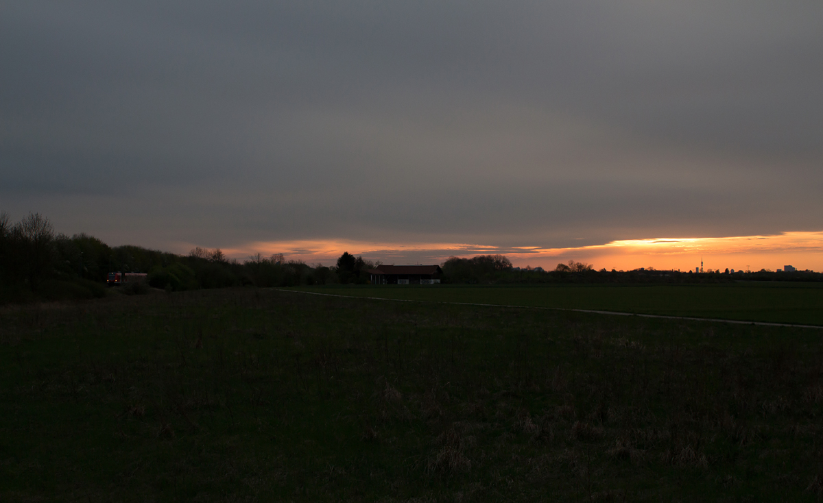 Feldkirchen und Umgebung eignet sich wahrlich kaum, um Eisenbahnbilder mit Motiv anzufertigen. Die Strecke ist bis auf wenige Ausnahmen uneinsichtig und dennoch fuhr ich einmal am Abend des 12. April 2017 in die  Pampa , da ich die Herausforderung liebe.

Anfangs, als die Sonne noch höher stand, herrschte ein völlig unattraktives Licht vor - eine große Wolkenfront verdeckte absehbar die letzten Sonnenstrahlen -
doch blickt man nach München mit seinem Olympiaturm, so gab es da noch ein letztes Wolkenloch, welches des nachts der Vergangenheit angehören wird.

Ein 423 als S2 in Richtung Erding wurde somit auserkohren, sich selbst mit der typischen Münchner Vorort-Landschaft und der bayrischen Landeshauptstadt im Bild zu vereinen.