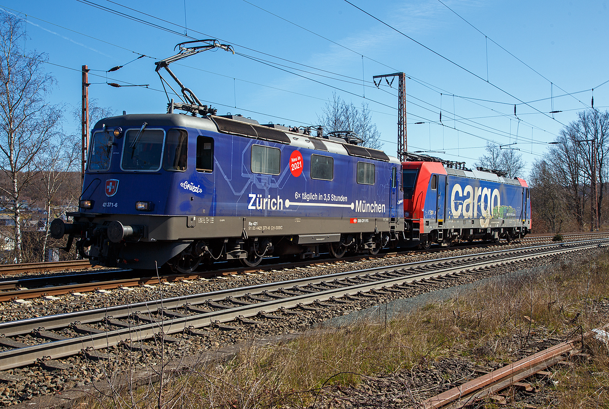 Ja, „Grezi“ oder „Servus“.....
Die SBB Cargo Re 421 371-6 (91 85 4421 371-6 CH-SBBC), ex Re 4/4 II 11371, mit Werbung Zrich – Mnchen 6 x tglich in 3,5 Stunden, fhrt am 30.03.2021 mit der SBB Cargo Re 484 016-1(91 85 4484 016-1 CH-SBBC), ex E 484.016 SR, einer TRAXX F140 MS am Haken durch Rudersdorf (Kr. Siegen), an der Dillstrecke (KBS 445), in Richtung Siegen.

Eine SBB Cargo Re 484 wie die 016-1 kann man eigentlich Deutschland nur mit viel Glck sehen, denn sie haben nur die Zulassungen fr die Schweiz und Italien. Sie haben keinen Stromabnehmer mit breiterer Wippe gem deutscher Vorschrift, auch die die deutsche Zugsicherung Indusi fehlt ihnen. Der Lz war auf dem weg nach Krefeld.

Lebenslufe:
Die Re 421 371-6 wurde 1983 von der SLM Winterthur (Elektrik BBC/ SAAS) unter der Fabriknummer 5235 gebaut und als Re 4/4 II 11371 an die SBB geliefert. Im Jahr 2002 erfolgte der Umbau zur Re 421 fr die SBB Cargo (Einsatz auch in Deutschland). Die zugelassene Hchstgeschwindigkeit fr die Schweiz betrgt 140 km/h und fr Deutschland 120 km/h.

Die Re 484 016-1 eine Bombardier TRAXX F140 MS wurde 2004 von Bombardier in Kassel unter der Fabriknummer 34009 gebaut und an die SBB Cargo AG, als E 484.016 SR geliefert. 