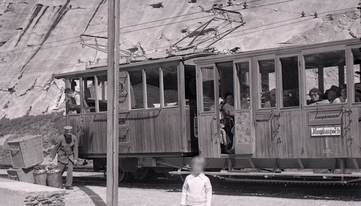 Jungfraubahn Lok 1 in Eigergletscher, zusammen mit dem Rowan-Wagen 1. Die Lok wurde 1898 abgeliefert und dann vor allem für den Tunnelbau eingesetzt. 1913 erhielt sie einen neuen Kasten. Es heisst, sie hätte 1951 wieder einen neuen Kasten bekommen. Ein Vergleich dieser Photo von 1955 mit dem jetzigen Zustand der Lok als Ausstellungsobjekt im Verkehrshaus Luzern (seit 1960) zeigt aber eine deutlich andere Fensteranordnung als 1955. Interessant an diesem Bild ist der - mit einem langen Aufenthalt verbundene - Güterumschlag in Eigergletscher. Zudem trägt das Personal der Jungfraubahn nicht das bei Schweizerbahnen übliche Dunkel, sondern eine graue Uniform. 