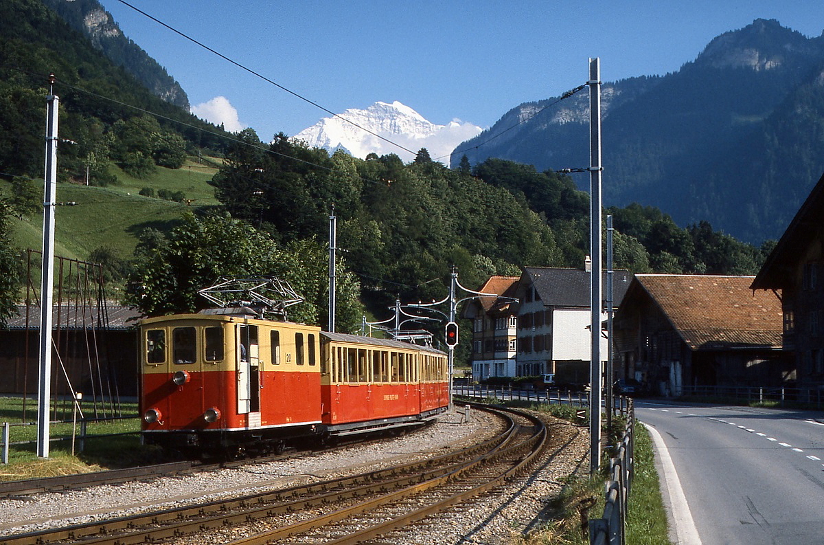 Kurz vor der Endstation in Wilderwil ist He 2/2 20 im Juni 1990 unterwegs. Im Vordergrund die Gleise der Berner Oberland-Bahn, im Hintergrund grüßt die 4.158 m hohe Jungfrau.