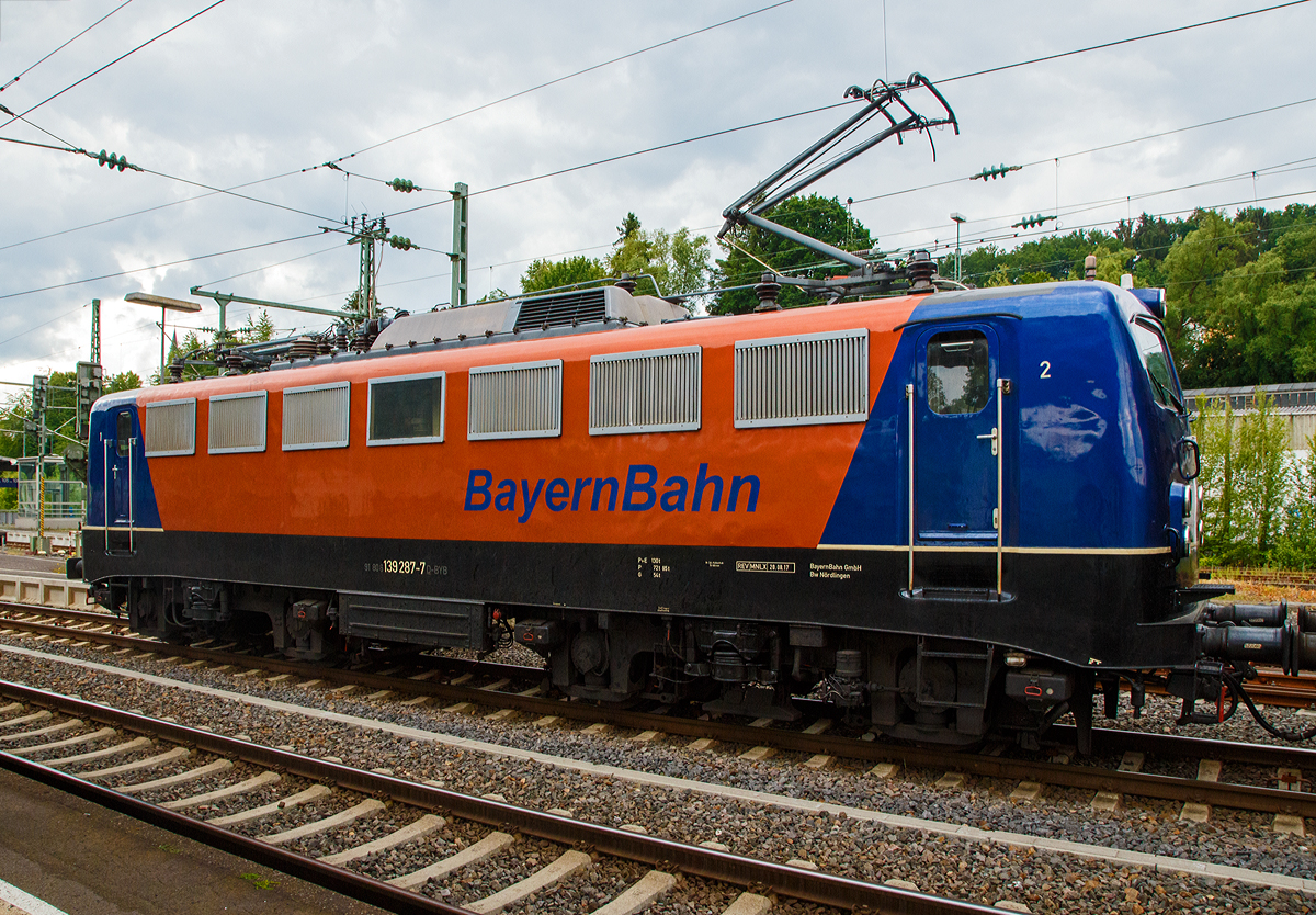 Noch ein Seitenblick.....
Die 139 287-7 (91 80 6139 287-7 D-BYB) der BayernBahn GmbH fhrt am 09.06.2020, mit dem sogenannten  Henkelzug  (Langenfeld/Rhld. nach Gunzenhausen), durch Betzdorf (Sieg) in Richtung Siegen. 	

Die Lok wurde 1963 von Krauss-Maffei AG in Mnchen-Allach unter der Fabriknummer 18956 gebaut, der elektrische Teil ist von den Siemens-Schuckert-Werke (SSW) in Berlin. Als DB E10 287 wurde in Dienst gesetzt, mit der Einfhrung des EDV-Nummernsystems wurde sie zum 01.01.1968 zur DB 110 287–0. Die Lok ist mit Einholmstromabnehmern ausgerstet, sie ist die letzte gebaute Kasten 110er. Zur 139er wurde sie erst 1994 nach einem Umbau, der Lokkasten wurde auf Drehgestelle der Baureihe 140 (E40) gesetzt und es folgte die Umzeichnung in DB 139 287-7. Zum Mai 2009 erfolgte die Z-Stellung bei der DB AG.

Zum 1.Oktober 2011 wurde die Lok durch die BayernBahn GmbH erworben und erhielt anschlieend eine Untersuchung. Die Neulackierung in kobalt-blau/orange erhielt sie dann 2017.
