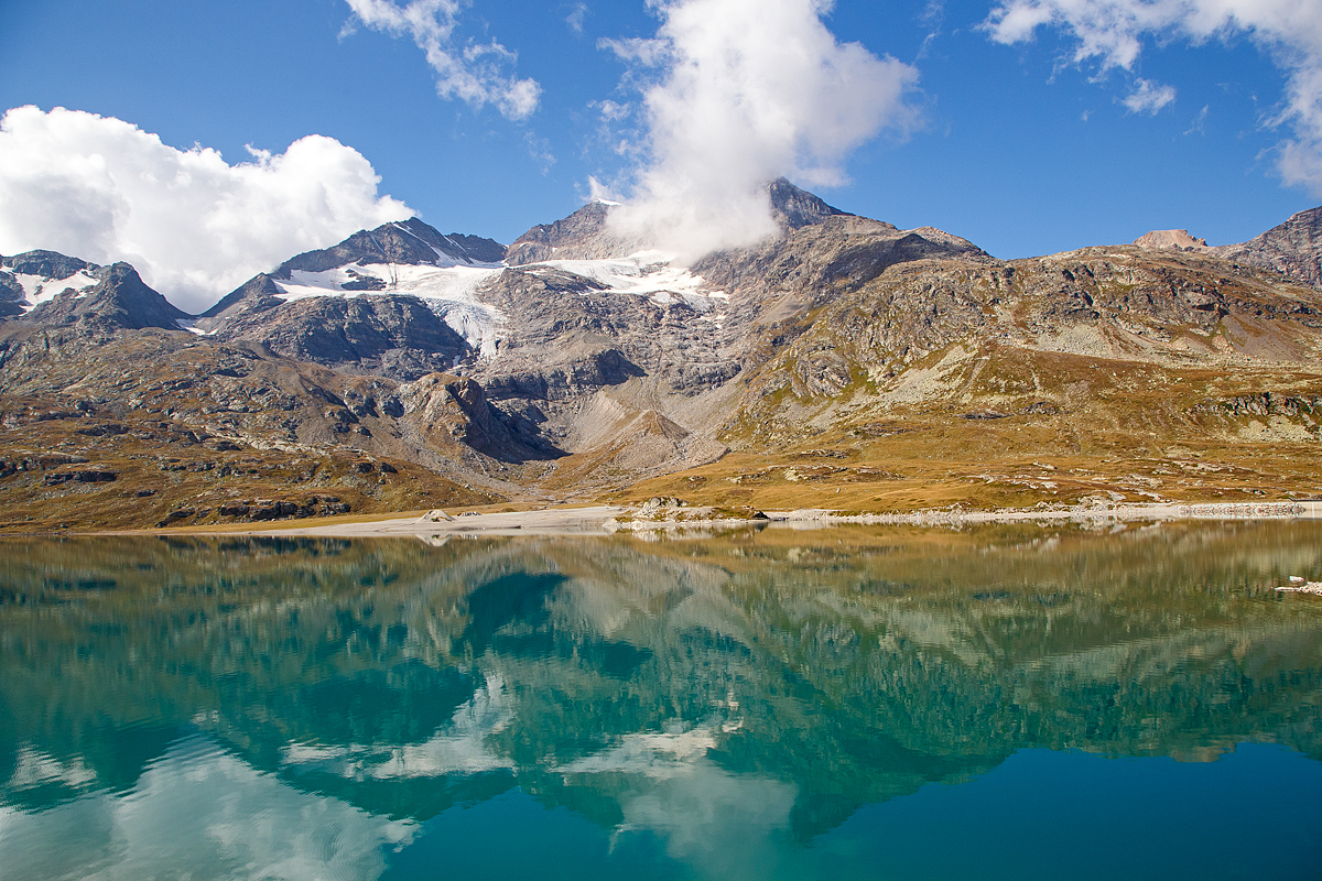 Nun erreichen wir am 06.09.2021den Lago Bianco auf einer Höhe von 2.223 Metern über Meer. 

Hier oben bei der Staumauer befindet sich auch zugleich die Wasserscheide zwischen Donau und Po. In Richtung Süden (Tirano) fließ das Wasser in den Po und somit ins Mittelmeer, in Richtung Norden (St. Moritz) fließ das Wasser über den Inn in die Donau und somit ins Schwarze Meer.

Der Lago Bianco ist ein Stausee am Berninapass zwischen dem Valposchiavo und dem obersten Seitental des Engadins, dem Val Bernina, in der Schweiz. Er liegt damit auf der Grenze zwischen den Bernina- (westlich) und Livigno-Alpen (östlich). Früher bestanden im Gebiet des heutigen Stausees zwei natürlichen Seen, der größere Lago Bianco, , und der kleinere südlich anschließende Lago della Scala. In den Lago Bianco mündeten mehrere kleine Bergbäche von den Höhen der Umgebung. Die beiden Gewichtsstaumauern Scala (Südseite) und Arlas (Nordseite) wurden zwischen 1910–1911 errichtet, so dass sich ein Stausee mit einem Volumen von 18,6 Mio. m³ bildete.

Unmittelbar am Nordende des Lago Bianco liegt die kontinentale Wasserscheide zwischen den Einzugsgebieten des Po und des Inn-Donau-Flusssystems. Wenige Meter nördlich des Lago Bianco liegt der Lago Nero (Schwarzsee) am Oberlauf des Berninabaches, dem folgt dann der kleinere Lago Pitschen (Pitschensee).