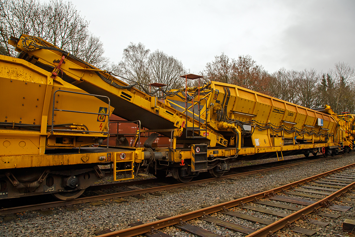 
Plasser & Theurer Material-, Förder- und Siloeinheiten MFS 38-D (teilweise auch BSW 2000 - Bunker-Schüttgut-Wagen genannt) der HERING Bahnbau, Burbach, Nr. 6 (Schweres Nebenfahrzeug Nr. D-HGUI 99 80 9552 125-3, ex 97 19 22 583 57-2, ist am 20.12.2020 auf dem Betriebshof der Westerwaldbahn (WEBA) auf der Bindweide bei Steinebach/Sieg abgestellt.

Diese MFS 38-D wurde 2002 von Plasser & Theurer gebaut. Die vierachsigen Fahrzeugrahmen und Drehgestelle sind moderne als die Wagen Nr. 1, 3 und 4 der Hering Bau.

Bei diesem MFS 38-D (das „D“ bedeutet Durchförderer) ist das Übergabeband starr (nicht schwenkbar), so ist ein  Entladen nebens Gleis nicht möglich. Die MSF 38-D haben ein Fassungsvermögen 38 m³ bzw. eine Nutzlast von 41,5 t, bei einem Eigengewicht von 38,5 t. Die Förderbänder werden hydraulisch angetrieben. Der Antrieb des Hydraulikaggregates erfolgt durch einen luftgekühlten Deutz Dieselmotor vom Typ F6L 912 mit einer Leistung von 82 kW.

TECHNISCHE DATEN:
Maschinenart: Material-Förder-Siloeinheit MSF 38
Hersteller: Plasser & Theurer
Spurweite: 1.435 mm (Normalspur)
Anzahl der Achsen: 4 (in 2 Drehgestellen)
Länge über Puffer:19.900 mm
Gesamtlänge mit Übergabeband: 23.550 mm
Drehzapfenabstand: 14.860 mm
Achsabstand im Drehgestell: 1.800 mm
Breite: 2.890 mm
Höhe: 4.280 mm
Eigengewicht: 38,5 t
Max. Zuladung (Nutzlast): 41,5 t
Ladevolumen: 38 m³
Höchstgeschwindigkeit: 100 km/h (geschleppt)
Kleister befahrbarer Gleisbogen: R 35 m
Bremse: KE – GP - A
Motor: luftgekühlter Deutz Dieselmotor vom Typ F6L 912 F
Leistung: 82 kW
Dieseltankvolumen: 750 l
Übergabeband: starr
Die Förderbänder sind in folgenden Geschwindigkeiten regelbar:
Siloband: 1. Gang (Speichern)   0 - 0,028 m/s / 2. Gang (Durchfördern)  0 - 0,140 m/s
Übergabeband: 0 - 1,000 m/s

Einsatz und Arbeitsweise:
Mit den Materialförder- und Siloeinheiten können unterschiedliche Schüttgüter wie beispielsweise Abraum, Planum- und Frostschutzmittel oder Bettungsstoffe unkompliziert, geladen, gefördert, gespeichert und entladen werden. Das Abraumverladesystem ist weitestgehend automatisiert und benötigt zur Bedienung nur wenig Personal. Das System verfügt zudem über eine autarke Energieversorgung (durch Dieselmotore). Ein unabhängiges Entladen ist durch schwenkbare Übergabebänder an jeder geeigneten Stelle möglich. Die MFS-Wagen können in Kombination mit einer Beladestation in beliebiger Anzahl aufgereiht und vor Kopf be- und entladen werden.

Am Boden jedes MFS 38 befindet sich ein über die ganze Wagenbreite gehender Fördergurt und an der Stirnseite ein breites Abgabeförderband. Das Aushubmaterial fällt von der Maschine kommend auf den Bodengurt, wird zum Abgabeförderband gebracht und an die nächste Einheit übergeben. Dieser Vorgang wiederholt sich, bis die am weitesten entfernte Siloeinheit erreicht wird. Bei dieser hintersten Siloeinheit ist das Abgabeförderband an der Stirnseite des Wagens in Ruhestellung. Auch der Fördergurt am Boden des Wagens ist so lange in Ruhestellung, bis die obere Füllgrenze erreicht ist. Dann wird der Fördergurt in Bewegung versetzt und dadurch im Übergabebereich freier Raum für die weitere Beladung geschaffen. Durch die dem Fülltempo angepasste Geschwindigkeit des Fördergurts wird die ganze Siloeinheit vollständig gefüllt. Das Fassungsvermögen einer Siloeinheit beträgt 38 Kubikmeter.

Nach Füllung einer Teilgruppe des Abraumzuges kann diese von der Restgruppe abgekuppelt werden, zu einer Entladestelle gefahren und dort entleert werden, während die Restgruppe des Abraumzuges noch befüllt wird. Das Entladeband ist nach beiden Seiten ausschwenkbar. Die Entladung kann dabei auf einer Deponie erfolgen oder es können LKW's oder Waggons am Nachbargleis beladen werden. Die MFS 38 (ohne nachfolgendes „D“) des Abraumzuges können durch Ausschwenken aller Abgabeförderbänder gleichzeitig entleert werden. Die Entladung eines Wagens dauert ungefähr 3,5 Minuten.