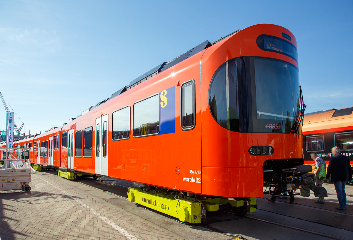 Stadler Rail präsentierte auf der InnoTrans 2018 (hier 18.09.2018) den neuen Worbla-Zug Be 4/10 für die RBS (Regionalverkehr Bern-Solothurn), hier in Form von dem RBS Be 4/10 Worbla 02. Bis 2020 sollen alle Mandarinli durch die Worblas bei der RBS ersetzt werden.
Der Triebzug steht hier auf Rollböcke der RailAdventure GmbH („Loco Buggies“).

RBS hat im Juni 2016 bei Stadler in Bussnang 14 vierteilige S-Bahn-Züge vom Typ Be 4/10 Worbla (Elektrischer Niederflurtriebzug) für die Linie S7 bestellt. Die vierteiligen elektrischen Meterspurzüge weisen 2 Triebköpfe am Ende und 3 Jakobs-Laufdrehgestelle als Verbindung zwischen den 4 Wagenkästen auf. Die Züge sind für eine maximale Geschwindigkeit von 100 km/h ausgelegt. Sie sind auf der gesamten Länge von 60 Metern durchgehend begehbar, was die Verteilung der Reisenden im Zug verbessert und das Sicherheitsgefühl erhöht. Die Triebzüge sind klimatisiert und erfüllen die Anforderungen des Behinderten-Gleichstellungsgesetzes. Niederflureinstiege und Schiebetritte zwischen Zug und Bahnsteig erleichtern den Eintritt. Modernste Technologien wie die voll redundante Traktionsanlage inklusive redundanter Leittechnik garantieren eine hohe betriebliche Verfügbarkeit und tiefe Life-Cycle-Kosten.

Technische Mekmale:
- Niederflurwagen in Alu-Leichtbauweise
- Luftgefederte Trieb- und Jakobslaufdrehgestelle
- Optimierte Motorleistung entsprechend des Einsatzes als Stadtbahn-Triebzug für den Betrieb auf Kurzstrecken
- Redundante Antriebsausrüstung (Antriebsstromrichter) in den Endwagen für hohe Ausfallsicherheit
- Acht Einstiegstüren für schnellen Fahrgastwechse
- Niederflureintritte an allen Türen
- Tür-Leuchtmelder (Countdown)
- Türsensor für «handsfree»-Eintritt
- Übersichtliche Plattformen
- Durchgehend begehbar
- Indirekte Beleuchtung
- Großzügige Multifunktionsabteile
- Klimaanlage

TECHNISCHE DATEN:
Bezeichnung: Be 4/10
Name: Worbla
Anzahl Fahrzeuge (geplant): 14
Spurweite: 1.000 mm (Meterspur)
Achsanordnung: Bo‘2‘2‘2‘Bo‘
Länge über Kupplung: 60.000 mm
Fahrzeugbreite: 2.650 mm
Fahrzeughöhe: 3.980 mm
Achsabstand in Drehgestell: 2.000 mm (Triebdrehgestell) / 2.150 mm (Laufdrehgestell)
Trieb- und Laufraddurchmesser: 770 mm (neu)
Höchstgeschwindigkeit: 100 km/h
Max. Leistung am Rad: 4 x 350 kW = 1.400 kW
Anfahrzugskraft: 120 kN (140 kN möglich)
Anfahrbeschleunigung: 1.0m/s² (1.2m/s² möglich)
Sitzplätze: 130
Stehplätze: max. 380
Fußbodenhöhe: 400 mm am Einstieg / 1.000 mm Hochflur
Einstiegbreite: 1 400 mm
Speisespannung: 1.250 V DC