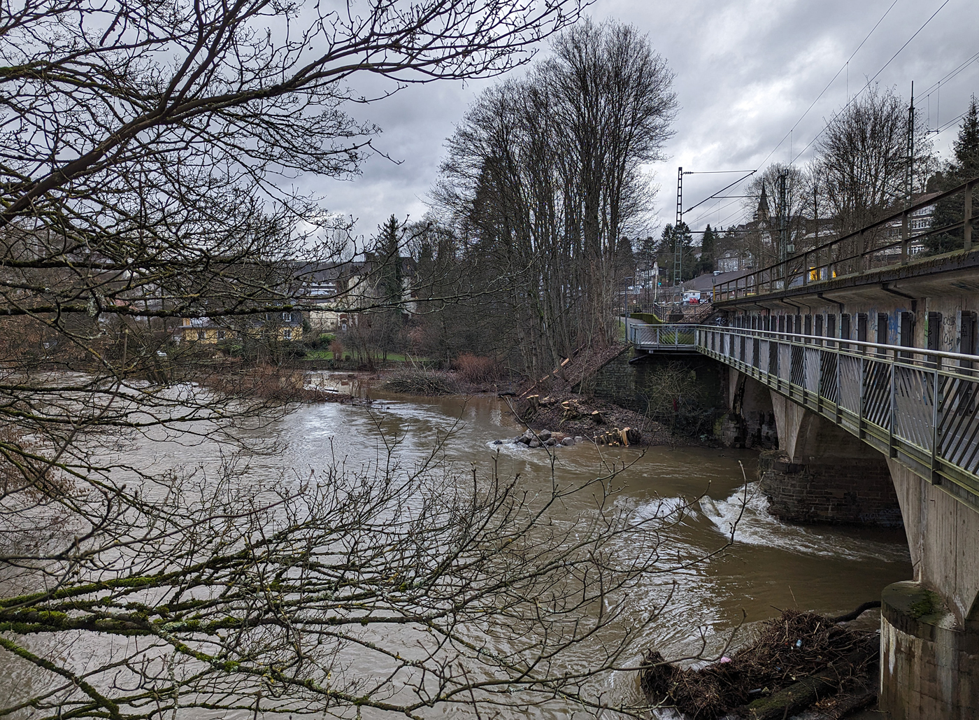 03.01.24	 Die vom Hochwasser beschädigte Eisenbahn-Siegbrücke mit Fußgängersteg in Kirchen (Sieg), der Siegstrecke (KBS 460), hier am 03.01.2024 (Bild mit Smartphone).

Die Bahnstrecke Siegen - Köln wird zwischen Kirchen und Betzdorf bis min. Ende nächster Woche gesperrt bleiben. Das hat die Deutsche Bahn Dienstagnachmittag mitgeteilt. Durch das Sieg-Hochwasser gibt es Schäden an einer Eisenbahnbrücke in Kirchen (kurz vorm Bahnhof). Aufgrund der Wetterlage und der weiterhin anhaltenden Regenfälle sei die Sperrung erforderlich, so die Bahn-Pressestelle. Die Züge des RE 9 können wegen der Streckensperrung nur bis Betzdorf fahren. Zwischen Siegen und Kirchen verkehrt die Regionalbahn 90 der Hessischen Landesbahn, leider aber nur wenige und teils mit „kleinen“ LINT 27 Triebwagen. Da wäre wahrhaft mehr möglich und man müsste den SEV nur zwischen Betzdorf und Kirchen betreiben. Aber aufgrund aktuell hoher Krankenstände kommt es auf der Linie RE 9 zudem zu Einschränkungen bis 12.01.2024.

Ich konnte mir den Schaden an einen Brückenpfeiler selbst anschauen (Bilder folgen) und finde die Streckensperrung richtig, darüber ließ ich auch keinen mit Personen besetzten Zug fahren. Was wäre wenn ein Zug darüber fährt und dadurch einbricht. Wer will das verantworten!!! 

Am 29.12.2023 wollten wir mit der Bahn nach Siegen, durch die Reiseauskunft unter www.bahn.de sahen wir bereits das durch wegen einer beschädigten Brücke zwischen Betzdorf (Sieg) und Kirchen (Sieg) ist eine Brücke beschädigt. Die Züge der Linie RE9 aus Richtung Köln enden und beginnen demnach in Betzdorf (Sieg). Die Züge der Linie RE 9 aus Richtung Siegen Hbf enden und beginnen demnach in Kirchen, diese waren aber nicht zu sehen. Bis dahin gab es keine Informationen zur Dauer der Sperrung. Somit fuhren wir mit dem Auto nach Kirchen und fuhren dann mit der RB 90 der HLB (Hessischen Landesbahn) in einem LINT 27, wie die Ölsardinen, nach Siegen Hbf. Auf der Rückfahrt um 14:31 Uhr (ab Siegen) mit RB 90 fuhren wir in einem LINT 41.

Am 30.12.2023 fuhren wir nochmal nach Siegen, das Wetter war besser und unser Ticket war noch gültig. Die Rückfahrt von Siegen machten wir um 13:31 Uhr mit dem HLB RB 90 nach Altenkirchen, dem HLB VT 261 einem LINT 41, dieser war der erste Zug der wieder durchfuhr und die Brücke in langsamer Fahrt befuhr. 

Später hieß es: Die Brücke zwischen Betzdorf und Kirchen ist nach wie vor beschädigt. Die Strecke ist aber wieder befahrbar. Die Züge fahren in dem betroffenen Streckenabschnitt langsamer. Reisende müssen mit Verzögerungen rechnen und sollten ihre Reiseverbindung kurz vor der Abfahrt des Zuges überprüfen.

Nun ist aber der betroffene Streckenabschnitt wieder gesperrt, die Brücke kann nicht mehr befahren werden.
 	google P6 pro						
