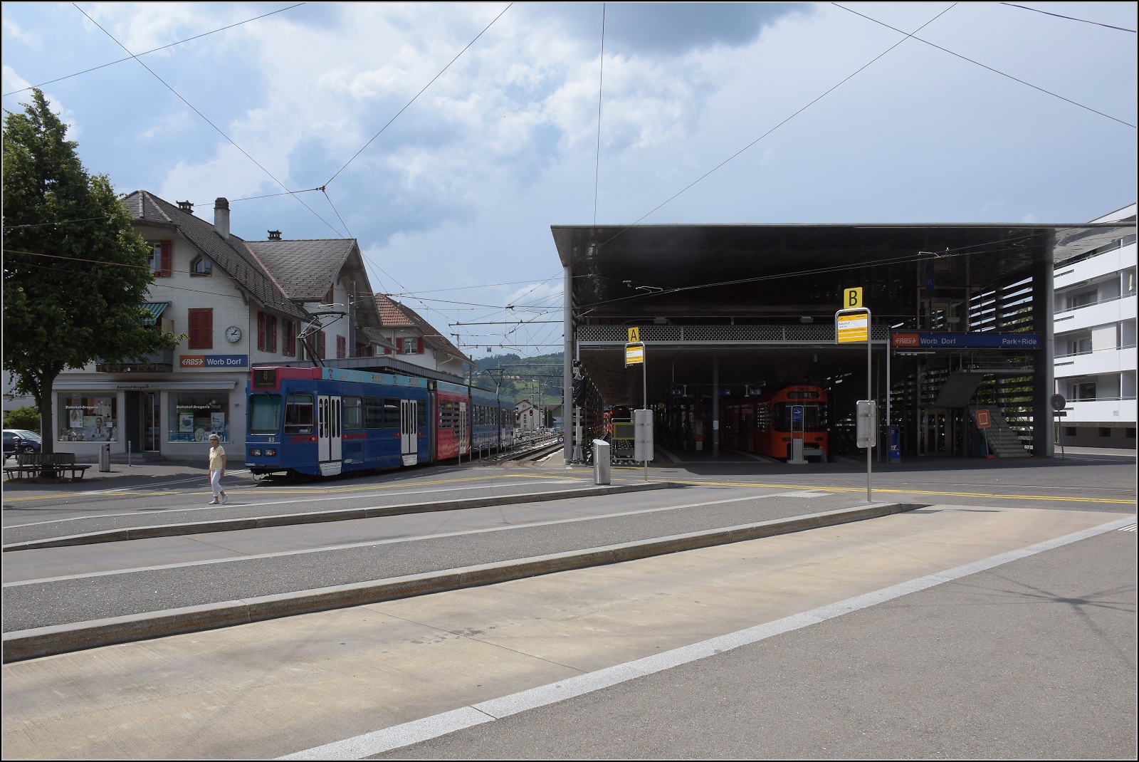 Bahnhof Worb links mit Haltestelle des Blauen Bähnli, heute Tramstrecke von Bernmobil, die Strecke aber im Besitz von RBS, rechts das Was-auch-immer-Gebäude mit der Worblentalbahn, wo sich zum Zeitpunkt des Fotos hinten das Blaue Bähnli versteckt. Vorne ist Tram 83 von Bernmobil als heutiges Blaues Bähnli im Bild. Juni 2023.