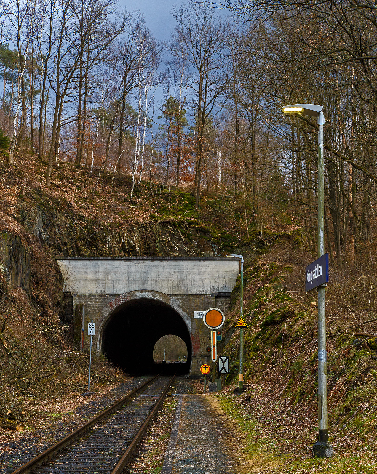 Der 137 m lange Herdorfer Tunnel hier am 18.03.2023 vom Hp Knigsstollen gesehen. 

Davor das Vorsignal (frs Einfahrtsignal Bf Herdorf) Vr 0 - Halt erwarten! Die gelbe Scheibe mit weiem Rand und gelbe Pfeil zeigt nach unten. Nachts leuchten auch die beiden gelben Lampen. Vor dem Signal die Ne 2 – Vorsignaltafel. Die Vorsignaltafel zeigt dem Lokfhrer, da das Signal, vor dem sie steht, Vorsignalfunktion hat, d.h., den Begriff eines nachfolgenden Hauptsignales vorankndigt. 
