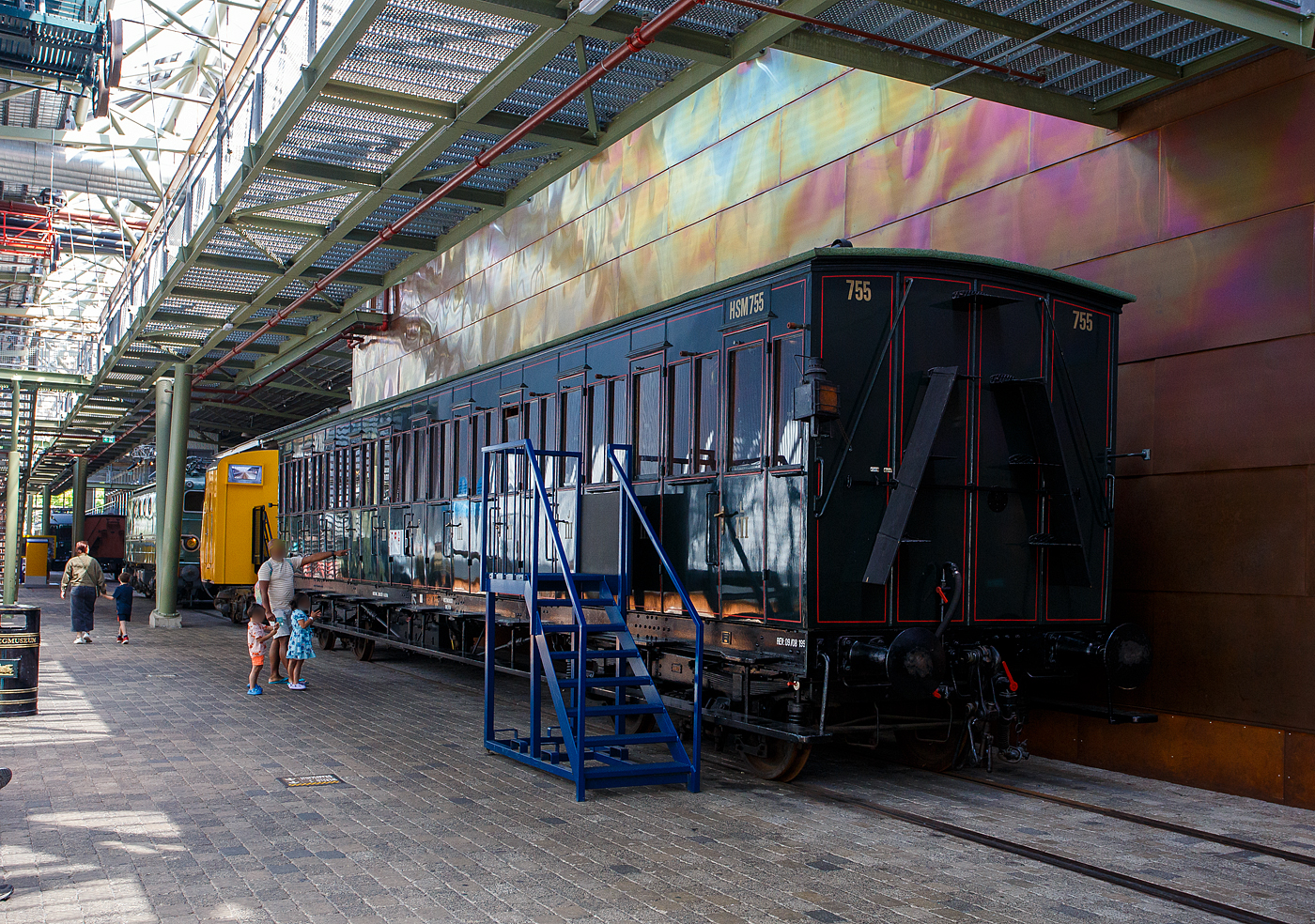 Der „viele Tren Wagen“ ein 3. Klasse Abteilwagen mit Seitentren ohne Durchgangsmglichkeit, HSM C 755 der damaligen HSM - Hollandsche IJzeren Spoorweg-Maatschappij (Hollndische Eisenbahn-Gesellschaft), ab 1922 NS C 5055, am 29 April 2025 im Het Spoorwegmuseum (bis 2005 NSM - Nederlands Spoorwegmuseum / Niederlndischen Eisenbahnmuseum) in Utrecht im ehemaligen Bahnhof Maliebaan.   

Ende des 19. Jahrhunderts hatten diese Wagen die Form eines langen, rechteckigen Kastens mit zahlreichen Tren an den Seiten, die Zugang zu den Sitzabteilen boten. Eine lange, auen angebrachte Stufe ermglichte dem Schaffner den bergang von einer Tr zur anderen. Die Sitzbereiche waren nur von auen zugnglich, einen Gang im Wageninneren gab es nicht. Anfang des 20. Jahrhunderts gab HSM 135 dieser Abteilwagen in Auftrag. Diese neue Wagengeneration war grer als die zuvor gebauten Wagen. Es gab 11 Abteile mit 10 Sitzpltzen fr 110 Personen. Dies brachte den Wagen den Spitznamen „Schafstall“ ein. Diese Wagen waren im ganzen Land bis in die 1950er Jahre im Einsatz, als neue Wagen und Triebwagen die veralteten Abteilwagen ersetzten.

Der Wagen vom Typ C11 (Serie 707 - 766 III) wurde 1907 von der Nederlandsche Fabriek van Werktuigen en Spoorwegmaterieel in Amsterdam (ab 1929 Werkspoor N.V.) gebaut und an die HSM geliefert.

Bedingt durch den Ersten Weltkrieg wurde 1917 eine Zweckgemeinschaft der unabhngigen Eisenbahngesellschaften HSM beziehungsweise HIJSM (Hollandsche IJzeren Spoorweg-Maatschappij) und SS (Maatschappij tot Exploitatie van Staatsspoorwegen) gegrndet. Die beiden Gesellschaften errichteten einen gemeinsamen Bahnbetrieb, blieben aber rechtlich unabhngig. Dies nderte sich 1938, als HIJSM und SS vollstndig fusionierten und in der AG NS (Nederlandse Spoorwegen, frher Nederlandsche Spoorwegen geschrieben) aufgingen. Das Rollmaterial wurde bereits 1921 zusammengelegt.

TECHNISCHE DATEN:
Spurweite: 1.435 mm (Normalspur)
Achsen: 4 (in 2 Drehgestellen)
Lnge ber Puffer: 18.300 mm
Leergewicht: 32.157 kg
Sitzpltze: 110 in der 3. Klasse in 11 Abteilen  10 Personen
Hchstgeschwindigkeit: 80 km/h

Quellen: Het Spoorwegmuseum, nmld.nl und Anschriften
Stand/berarbeitung: September 2025
