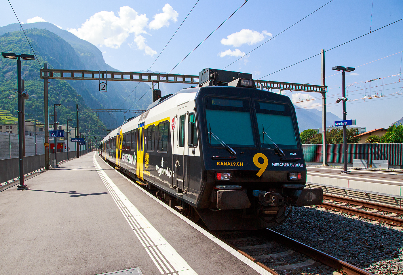 Der dreiteilige NPZ Domino RA14 der RegionAlps, mit der Vollwerbung für den Walliser Fernseh-Sender Kanal 9, verlässt am 26.Mai 2023, als Regionalzug nach Monthey den Bahnhof Martigny.

Der NPZ Domino besteht aus den Triebwagen RBDe 560 DO RA 94 85 7 560 414-5, dem 2.Klasse Wagen B NPZ DO RA 50 85 29-43 414-6 und dem 1./2. Klasse Steuerwagen ABt NPZ DO RA 50 85 39-43 954-9 CH-RA (ex SBB RBDe 560 276, ex SBB B 29-43 181 und ex SBB ABt 39-43 869).

Die Regionalps (RA) ist ein Eisenbahnverkehrsunternehmen im Kanton Wallis, mit Sitz in Martigny hat. Eigentümer derim Jahr 2003 gegründete Gesellschaft  sind die Schweizerischen Bundesbahnen (SBB) mit 70 %, die Transports de Martigny et Régions (TMR) mit 18 % und der Kanton Wallis mit 12 %. Die RA betreibt den Regionalverkehr vor allem im Rhonetal zwischen dem Ostufer des Genfersees und Brig. Inzwischen gehören Regionalps 16 dreiteilige NPZ Domino (Trieb-, Zwischen- und Steuerwagen ex SBB) und 4 RABe 525 NINA (ex TMR).
