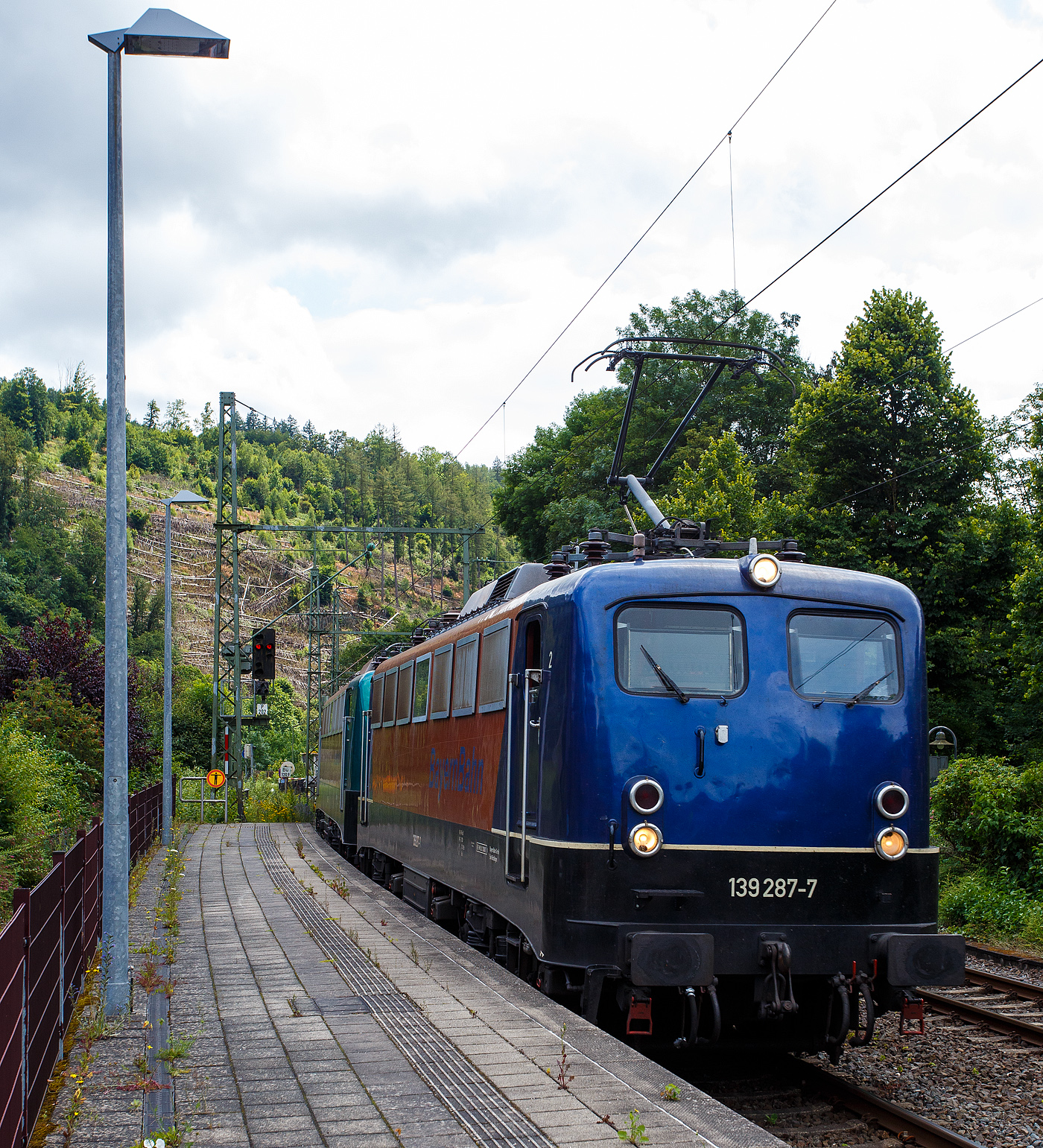 Die 139 287-7 (91 80 6139 287-7 D-BYB) mit der kalten 140 850-9 (91 80 6140 850-9 D-BYB) beide von der BayernBahn GmbH fahren am 04 Juli 2024 mit dem sogenannten  Henkelzug  (Langenfeld/Rhld. nach Gunzenhausen), durch den Bahnhof Kirchen (Sieg) in Richtung Siegen.

Die 139 287-7:
Vorne die 139 287-7 wurde 1963 von Krauss-Maffei AG in München-Allach unter der Fabriknummer 18956 gebaut, er elektrische Teil ist von den Siemens-Schuckert-Werke (SSW) in Berlin. Als DB E10 287 wurde in Dienst gesetzt, mit der Einführung des EDV-Nummernsystems wurde sie zum 01.01.1968 zur DB 110 287–0. Die Lok ist mit Einholmstromabnehmern ausgerüstet, sie ist die letzte gebaute Kasten 110er. Zur 139er wurde sie erst 1994 nach einem Umbau, der Lokkasten wurde auf Drehgestelle der Baureihe 140 (E40) gesetzt und es folgte die Umzeichnung in DB 139 287-7. Zum Mai 2009 erfolgte die Z-Stellung bei der DB AG. Zum 1.Oktober 2011 wurde die Lok durch die BayernBahn GmbH erworben und erhielt anschließend eine Untersuchung. Die Neulackierung in kobalt-blau/orange erhielt sie dann 2017.

Die 140 850-9
Die (E40) 140 850-9 wurde 1972 bei Henschel & Sohn in Kassel unter der Fabriknummer 31696 gebaut, der elektrische Teil wurde von der AEG in Berlin unter der Fabriknummer 8804 geliefert/gebaut. Als 140 850-9 wurde sie an die Deutsche Bundesbahn ausgeliefert, die Ausmusterung bei der DB Cargo AG erfolgte im Juli 2017 und sie wurde an die BayernBahn GmbH in Nördlingen verkauft.