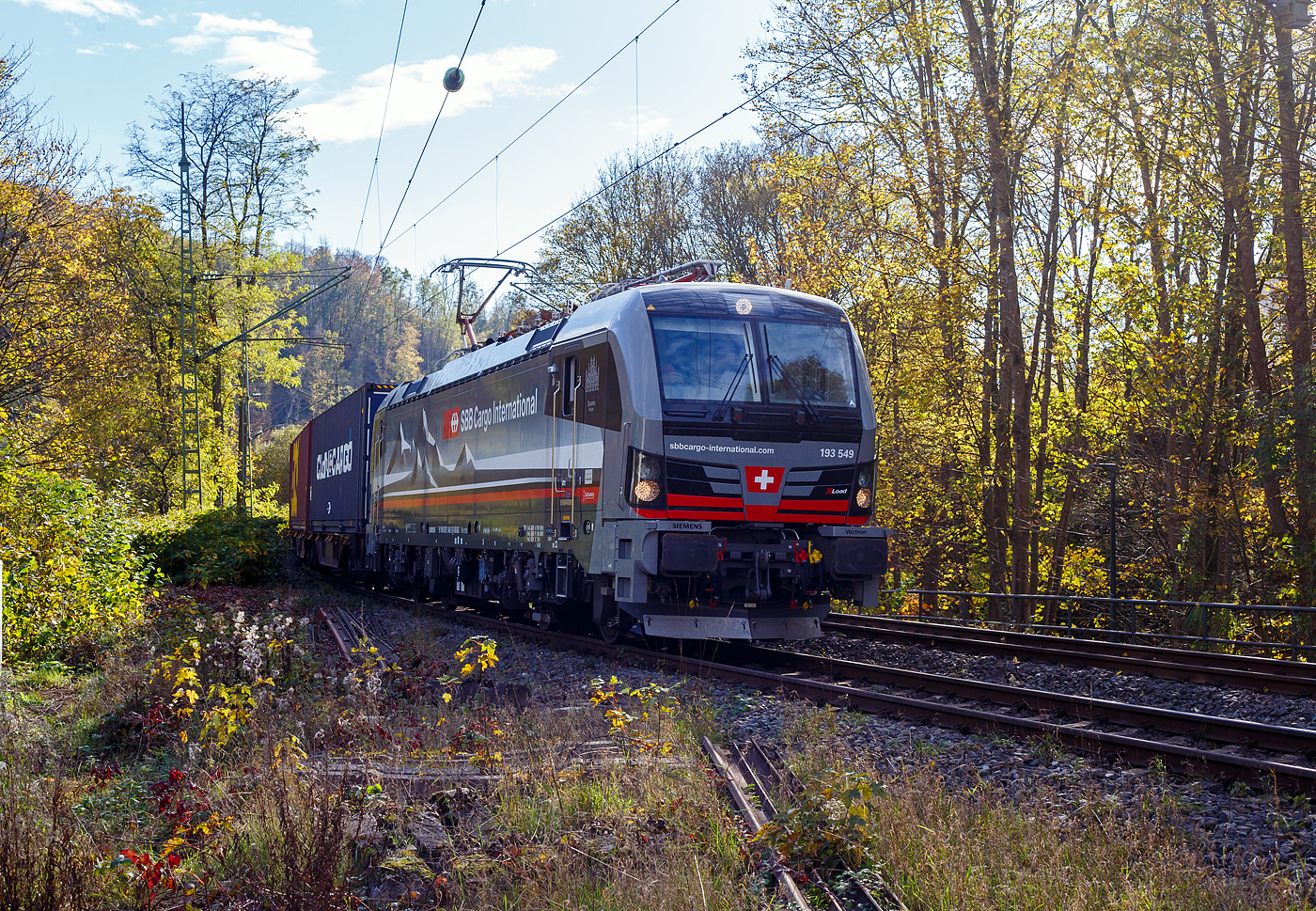 Ein fast fabrikneuer SIEMENS XLoad-Vectron, gerade mal 4 Wochen alt.
Die an die SBB Cargo International AG vermietete 193 539 mit XLoad  Duomo Milano  (91 80 6193 549-3 D-SIEAG) der SüdLeasing GmbH, Stuttgart (eingestellt in Deutschland durch Siemens) fährt am 29.10.2024 mit einem KLV-Zug, durch Kirchen (Sieg) in Richtung Siegen.

Die Multisystemlokomotive Siemens Vectron MS wurde 2024 von Siemens Mobilitiy in München-Allach unter der Fabriknummer 23595 gebaut und am 25.09.2024 ausgeliefert. Sie wurde in der Variante A40-1a ausgeführt und hat so die Zulassung für Deutschland, Österreich, die Schweiz, Italien, die Niederlande und Belgien (D / A / CH / I / NL / B). Sie verfügt über eine Leistung von 6,4 MW (160 km/h) und ist neben den nationalen Zugsicherungssystemen mit dem Europäischen Zugsicherungssystem (ETCS  BL3) ausgestattet. Zudem ist sie mit der neuen Ausrüstungspaket XLoad ausgestattet. 

Das neue XLoad Ausrüstungspaket für Vectron:
XLoad ist ein Ausrüstungspaket für Vectron, welches künftig mitbestellt, aber auch bei bereits ausgelieferten Vectron Loks nachgerüstet werden kann. Das Feature verbessert die Reibwertausnutzung und ermöglicht dadurch höhere Anhängelasten. Zudem reduzieren die Fahreigenschaften, die das Feature bewirken, den Verschleiß von Rad und Schiene.

Aktuell sind die Schweizer Vectron-Lokomotiven (SBB Cargo und BLS Cargo) in der Regel in Doppeltraktion unterwegs. Die Steigungen und Rampen der Schweizer Berge sind vor allem bei schlechten Witterungsbedingungen nicht ohne. Eine Lokomotive muss auch bei geringerem Schlupf genügend Traktion auf die Schienen bringen, um alle Güter sicher und zuverlässig ans Ziel zu bringen. Ein effizienter Weg aus dieser «Misere» ist die für Vectron entwickelte Zusatz-Funktion «XLoad». Den erfolgreichen Beweis trat eine SIEMENS Testlokomotive im Frühjahr 2022 bei der SBB Cargo International und bei der BLS Cargo eindrücklich an. 

Für SBB Cargo International bewies die Test-Lokomotive am Bözberg und für BLS Cargo an der Nordrampe des Lötschbergs ihre enorme Zugkraft. 
Vectron meisterte im Frühjahr 2022 die lange 12‰-Steigung des Bözbergs mit einer Anhängerlast von 2.000 Tonnen bravourös. Bei den nächtlichen Testfahrten zeigte sich eindrücklich die enorme Zugkraft der Lokomotive. 

Am Lötschberg wurden bei der BLS Cargo steigungsmäßig noch ein paar Promille draufgepackt. Mit 1.020 Tonnen im Gepäck bewältigte die Vectron-Lokomotive mit XLoad-Feature die 27‰-Steigung der Nordrampe ebenfalls meisterlich. Und auch diverse Anfahrtsversuche absolvierte der mit dem XLoad-Feature aufgerüstete Vectron problemlos. 

So bestellte die SüdLeasing GmbH (Stuttgart) im Auftrag der SBB Cargo International jüngst 20 Vectron Lokomotiven mit XLoad bei SIEMENS.
