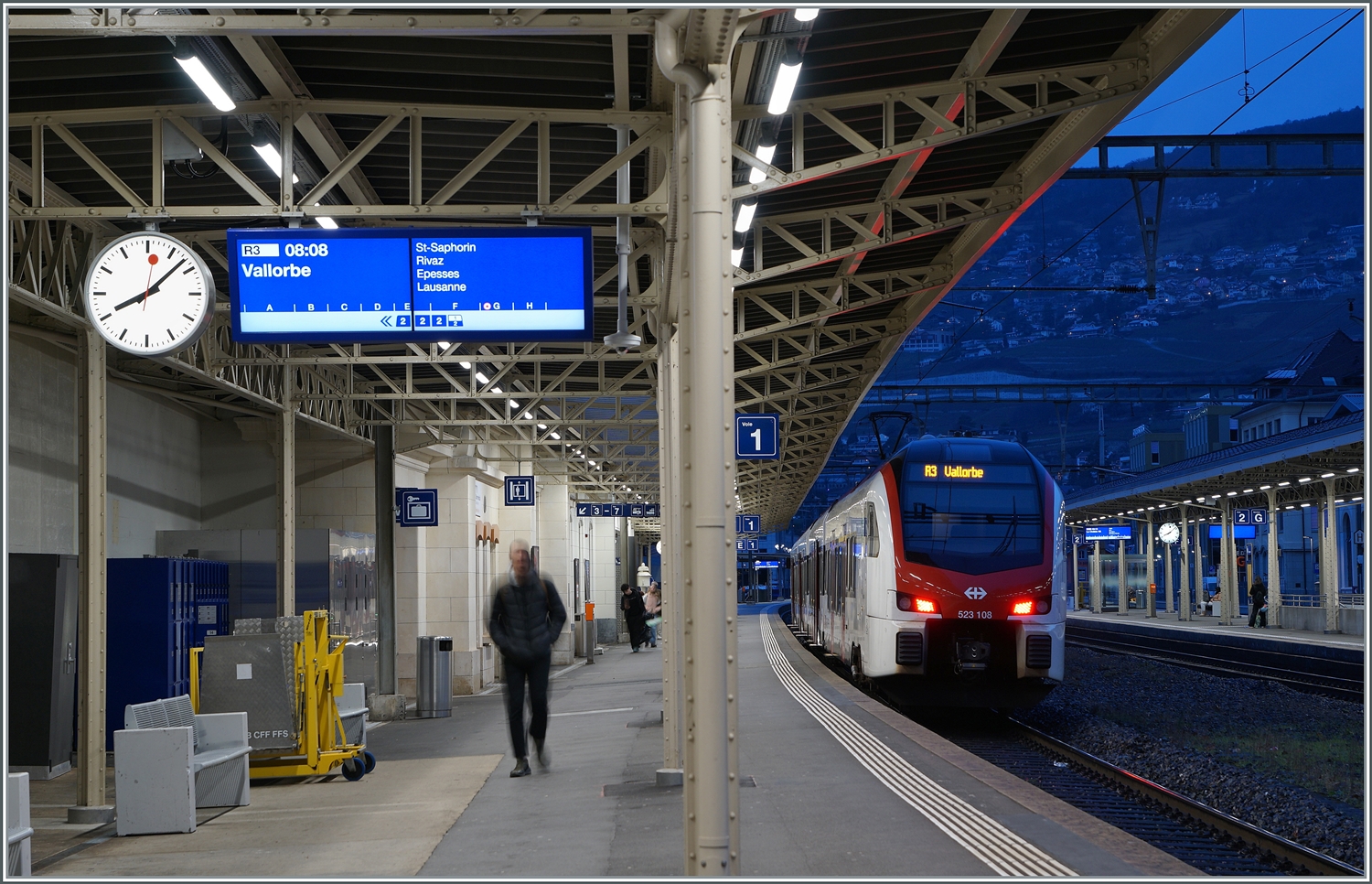 Ein Vorteil der Fotografie in der Blauen Stunde mit langer Belichtungszeit ist, dass Menschen in Bewegung unscharf werden und somit nicht erkannt werden. In Vevey zeigt sich der SBB RABe 523 108 als R3 nach Vallorbe. Zum Fahrplanwechsel wurde die  S-Bahn  im Raum Lausanne wieder durch  Regio -Züge ersetzt, eigentlich sinnvoll den S-Bahn verstand hier ohnehin niemand. 2. Januar 2024