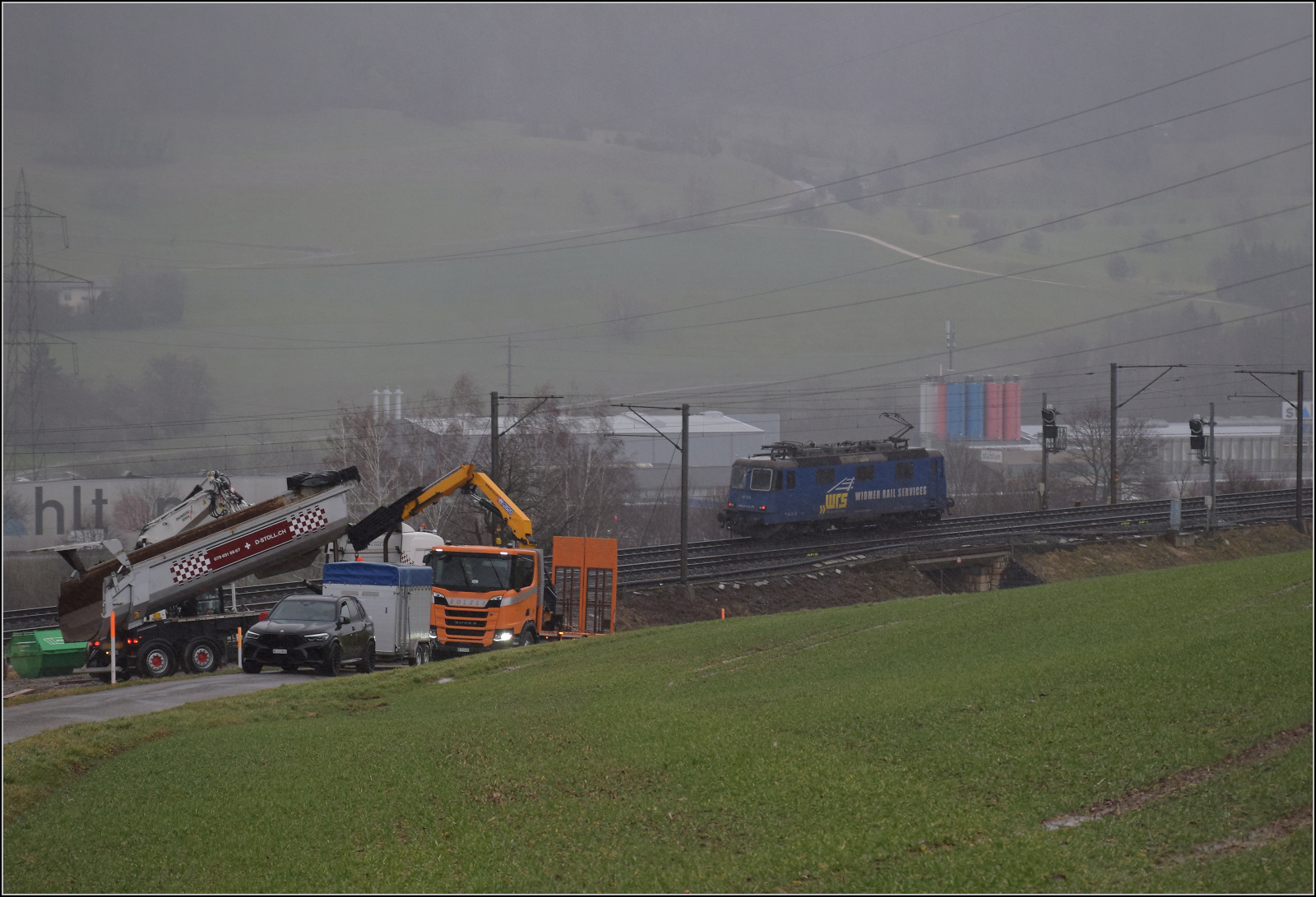 Schlechtwetter und ein Baustellendrama bei Frick. 

Re 421 373 der WRS ist solo auf dem Weg zum B�zberg. Dabei kommt die Lok an einer veritablen Havariestelle vorbei. Denn bei der ung�nstigen Abschleppaktion mit dem ge�ffneten Muldenkipper kam die Sache ruckartig in Bewegung, wegen der Schr�glage spritzte pl�tzlich das Hydraulik�l in einer Wolke aus dem Hydraulikzylinder, die Mulde rauschte nach unten. Ein Knall und der Schlag des Aufpralls der Mulde auf den Sattel des LKWs kamen kurze Zeit sp�ter an. Mein ungutes Gef�hl, die werden doch nicht..., hat sich best�tigt. Schr�glage und eine ruckartige Befreiung aus den selbstgegrabenen L�chern sind eben doch zu viel, selbst f�r die beste Hebehydraulik. F�r die niedrige Bahnbr�cke mussten nun die noch verbliebenen Hydraulikzylinder noch aus dem Weg und die festsitzende Heckklappe runtergeklappt werden. Anschlie�end die instabile Mulde gesichert werden. Und wir sitzen auch im Superschlechtwetter fest. Februar 2026.