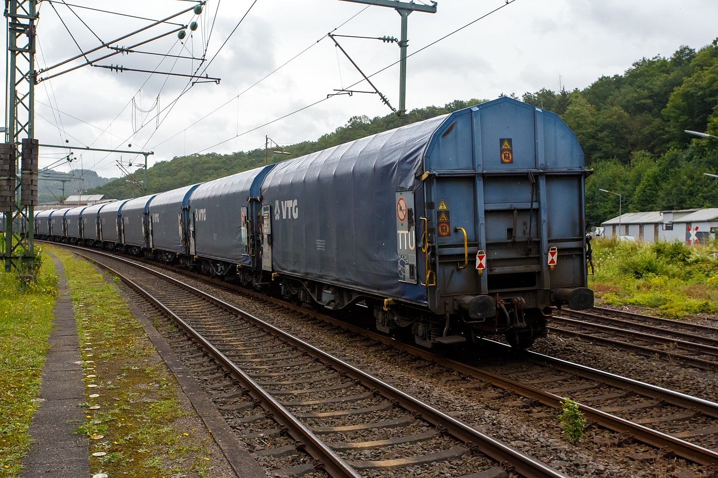 Und tschüss bis gleich in Herdorf, der vierachsiger Drehgestell-Flachwagen mit verschiebbarem Planenverdeck und Lademulden für Coiltransporte, 37 80 4668 122-7 D-VTG, der Gattung Shimmns-ttu (VTG Bez. F46.520D), der VTG Rail Europe GmbH (Hamburg), am 03 Juli 2024 eigereiht im Zugverband (Zudschluss) der KSW bei einer in Zugdurchfahrt im Bf Scheuerfeld/Sieg, in Richtung Herdorf via Betzdorf.

Diese Wagen sind speziell für den Transport witterungsempfindlicher Blechrollen (Coils) bestimmt. Er besitzt feste Stirnwände, 5 fest im Untergestell eingebaute Lademulden. Die Wagenabdeckung erfolgt mit einer PVC-beschichteten Gewebeplane, die auf 11 Rohrrahmen aufliegt. Über Laufwagen ist das Planendach in Wagenlängsrichtung verschiebbar. Die Verriegelung des Planendaches an den Stirnwänden erfolgt über eine 4-Punkt-Zentralverriegelung, die sowohl vom Erdboden als auch von der Rampe aus bedient werden kann. Das Planenverdeck lässt sich an einem Wagenende so weit zusammenschieben, dass ca. 2/3 der Ladefläche zur Beladung freigegeben wird. Hierdurch ist eine einfache Beladung des Wagens sowohl von oben als auch von der Seite aus möglich.

TECHNISCHE DATEN:
Spurweite: 1.435 mm
Achsanzahl: 4 (in 2 Drehgestelle)
Länge über Puffer: 12.040 mm
Länge der Ladefläche: 10.800 mm
Drehzapfenabstand: 7.000 mm
Achsabstand im Drehgestell: 1.800 mm
Eigengewicht: 22.250 kg
Höchstgeschwindigkeit: 100 km/h (beladen) / 120 km/h (leer)
Maximales Ladegewicht: 67,7 t (ab Streckenklasse D4), DB CM 61,7 t
Mulden: 5 Stück, im Untergestell integriert
Werkstoff Muldenmatten: Tecthan
Festlegearme: 20 Stück, stufenlos verstellbar, ausgestattet mit Schutzplatten
Coil-Breite: min. 342 mm bis max.  2.042 mm
Kleinster bef. Gleisbogenradius: 35 m
Bauart der Bremse: KE-GP-A (K)
Bremssohle: C 810
Intern. Verwendungsfähigkeit: TEN-GE