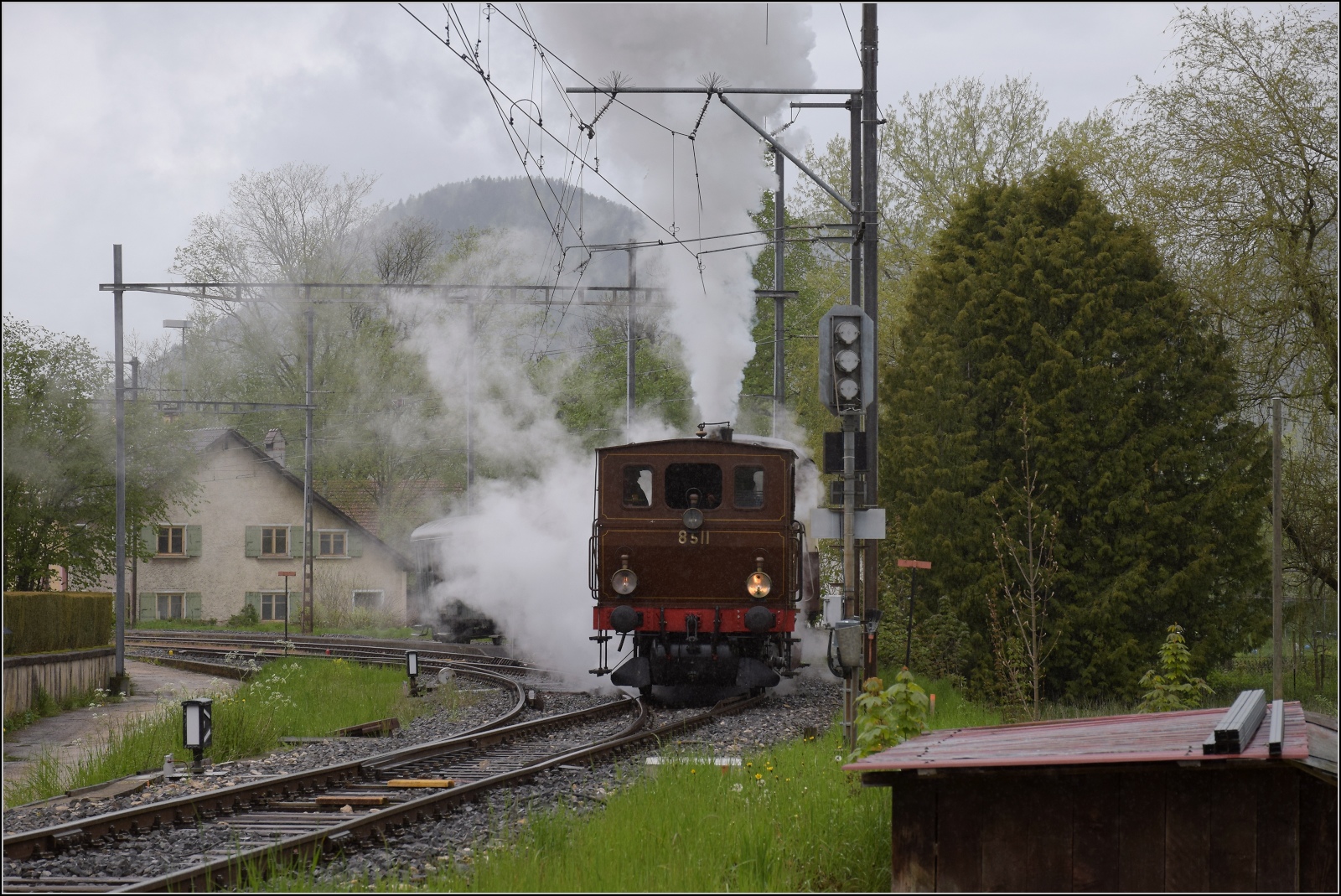 Vapeur Val-de-Travers: Train  Au fil de l'Areuse .

E 3/3 8511 steht in der Bahnhofsausfahrt von Môtiers -  Bahnübergangsstörung, für ein Bild mit Münster ist der Zug leider gerade nicht weit genug vorgefahren. Mai 2023.