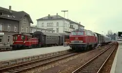 215 005-0 wartet um 1980 vor einem Eilzug nach Ulm im Bahnhof Friedrichshafen Stadt auf Fahrg�ste, der Dampf zwischen Lok und Packwagen zeigt, dass die Dampfheizung in Betrieb ist.